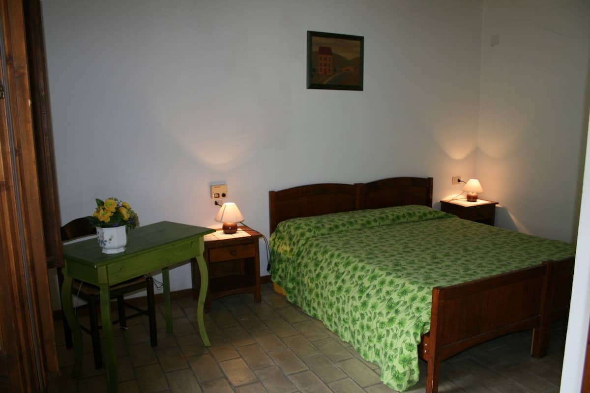 A small, neatly arranged bedroom with a single bed, a wooden nightstand, a desk with a chair, and a wall-mounted lamp.