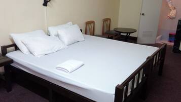Una cama individual con ropa de cama blanca, una mesita de noche, una silla, una pequeña mesa y una puerta con un abrigo colgado.