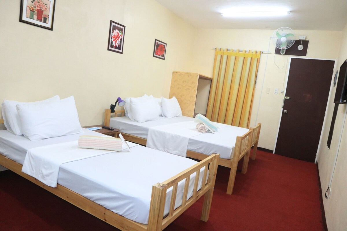 Une chambre avec deux lits simples, un bureau avec une chaise, une lampe de bureau et un ventilateur fixé au mur.