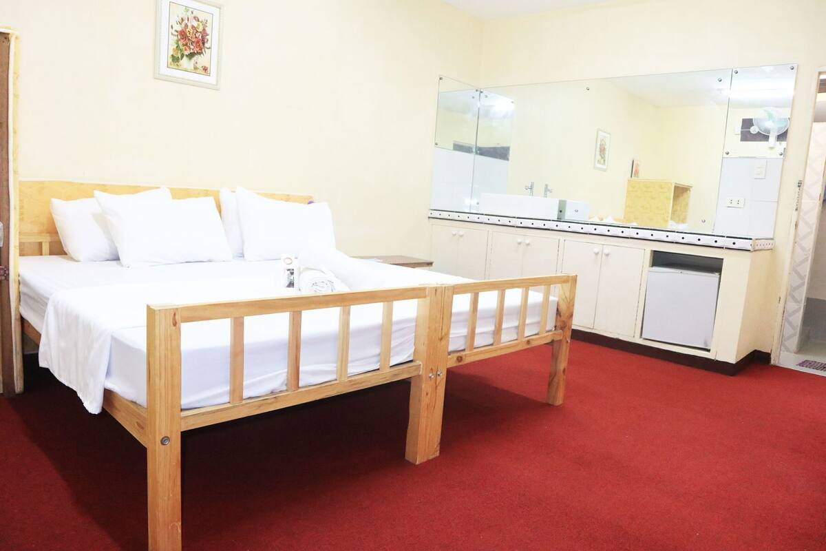 Una habitación con una cama de madera, alfombra roja, baño blanco con un espejo grande y un armario pequeño.