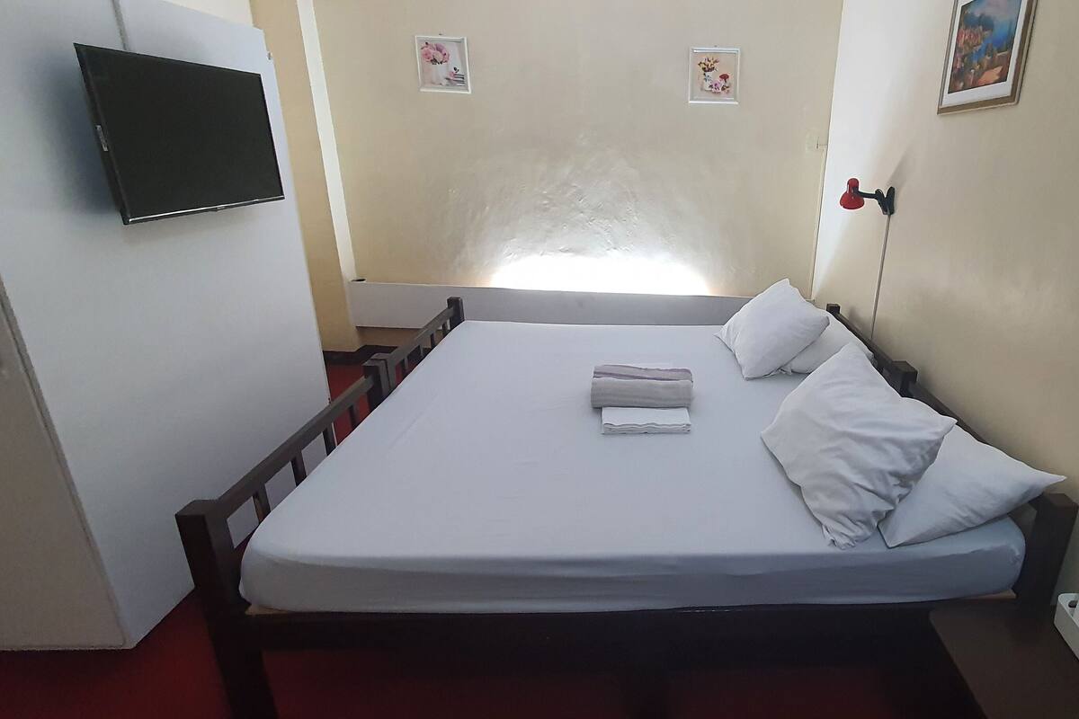 Una habitación de hotel con una cama, un televisor montado en la pared, una lámpara de noche y un teléfono también en la pared.