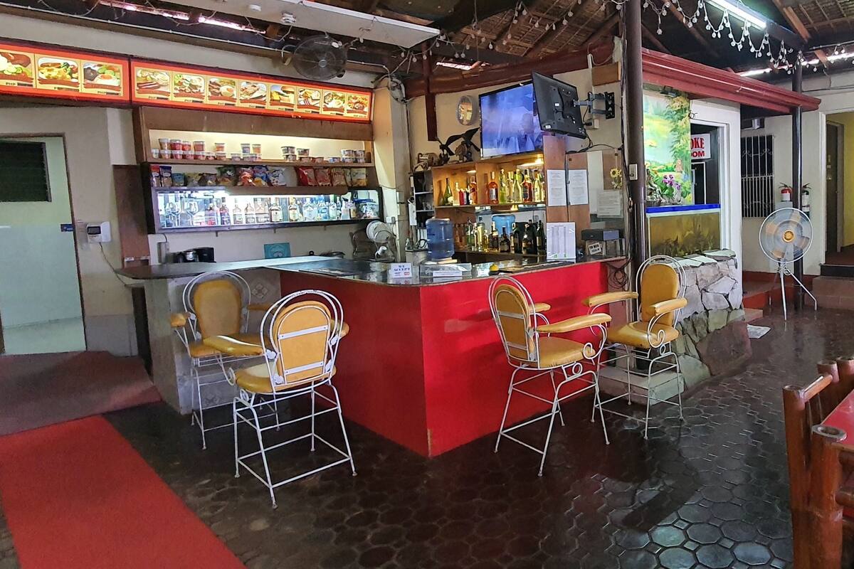 Zona de bar con una barra roja, diversas opciones de asientos incluyendo taburetes y sillas, un televisor montado en la pared y estanterías llenas de bebidas.