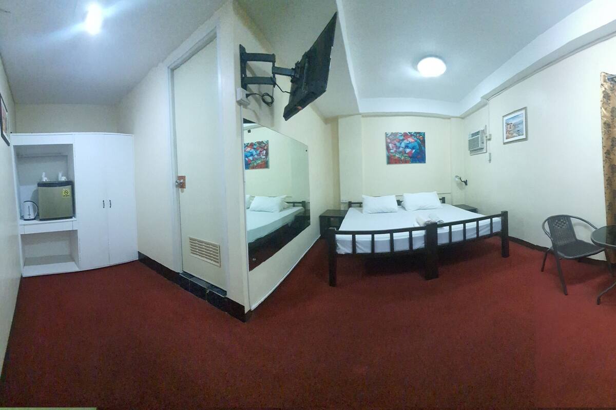 Habitación de hotel con cama, una mesita con dos sillas, espejo, televisor montado en la pared y ventana con cortinas.