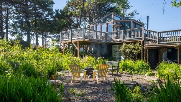 A cabin with a deck, surrounded by lush greenery and tall trees. - Zen Beach Retreat-Relaxing Sauna - Steps to a Sandy Beach! (Yachats)