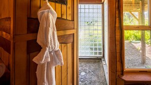 A wooden interior with a white robe hanging on a rack, a glass door leading outside, and a view of greenery. - Zen Beach Retreat-Relaxing Sauna - Steps to a Sandy Beach! (Yachats)