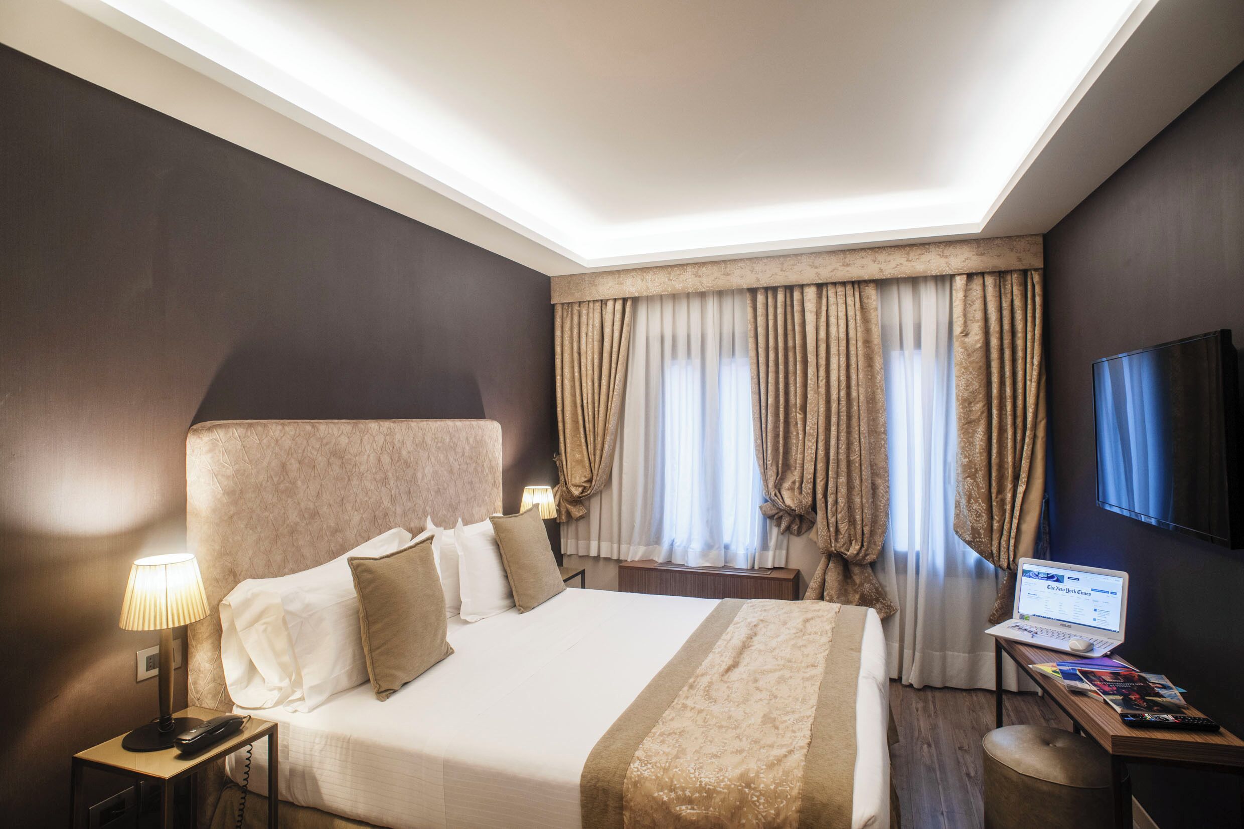 Une chambre d’hôtel avec un grand lit, un bureau avec un ordinateur portable, une télévision à écran plat et une table de chevet avec une lampe et un téléphone.