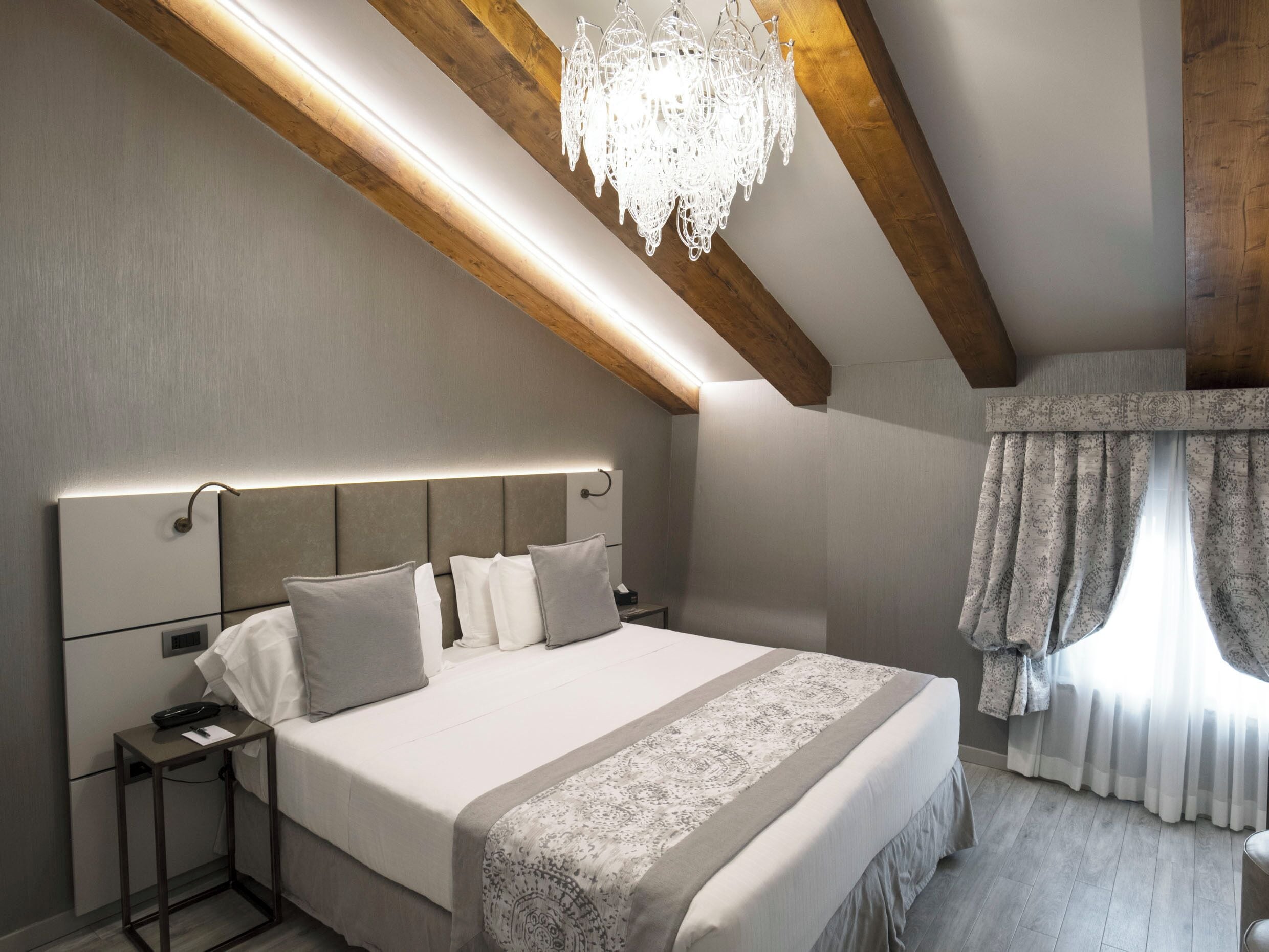 A bedroom with a large bed, a bedside table, a nightstand, a chandelier, and a window with curtains.