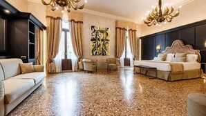 A spacious room with a large bed, a sofa, a coffee table, and two armchairs. There are two large windows with curtains, a chandelier, and a painting on the wall. - Hotel Aquarius Venice (Venice)