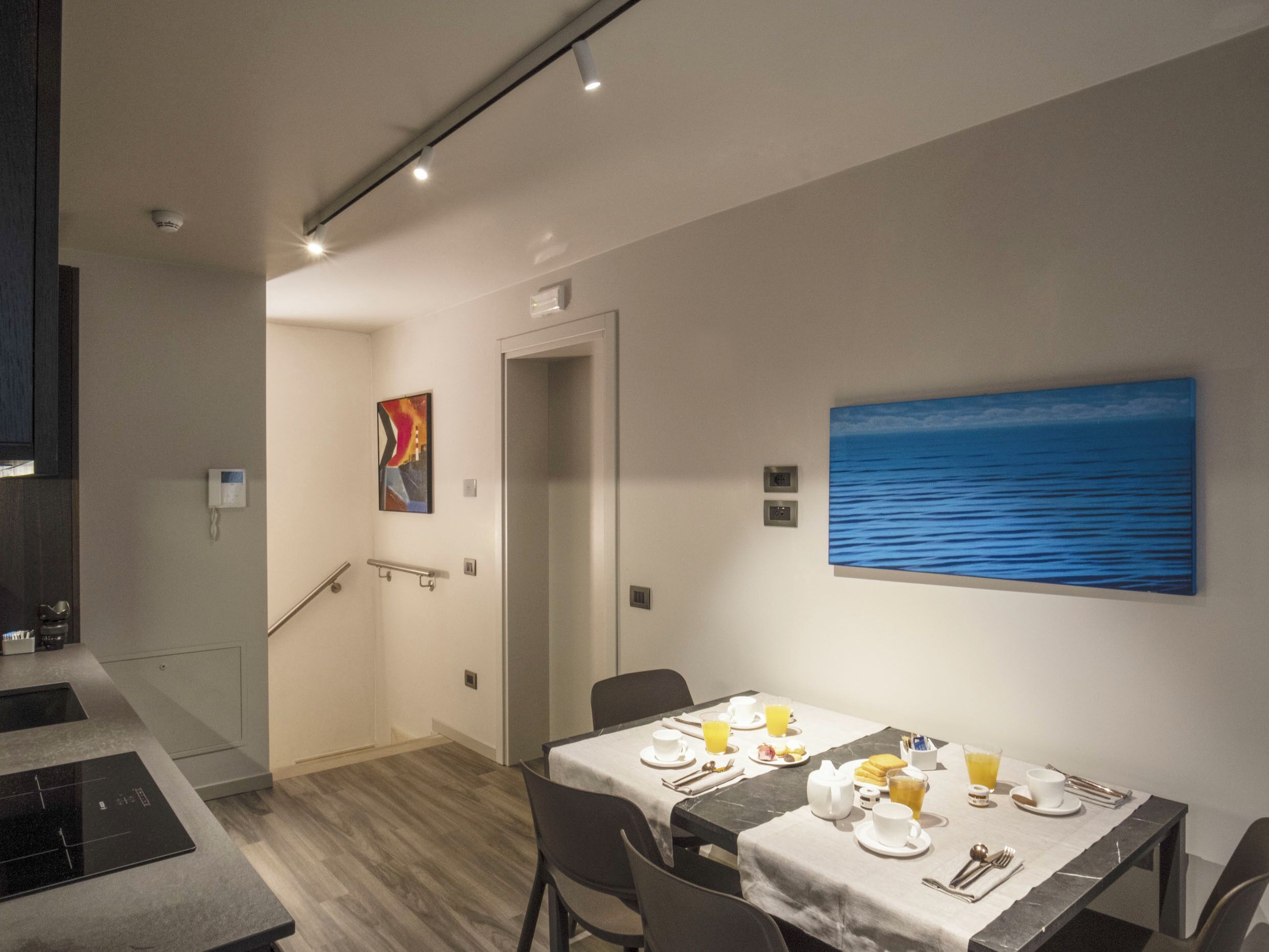 Un comedor moderno con una mesa dispuesta para el desayuno, un cuadro del océano en la pared y una escalera que lleva a otra habitación.