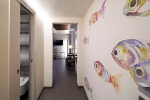 Corridoio d'hotel con un bagno a sinistra, una zona salotto al centro e una parete decorata con opere d'arte raffiguranti pesci colorati.