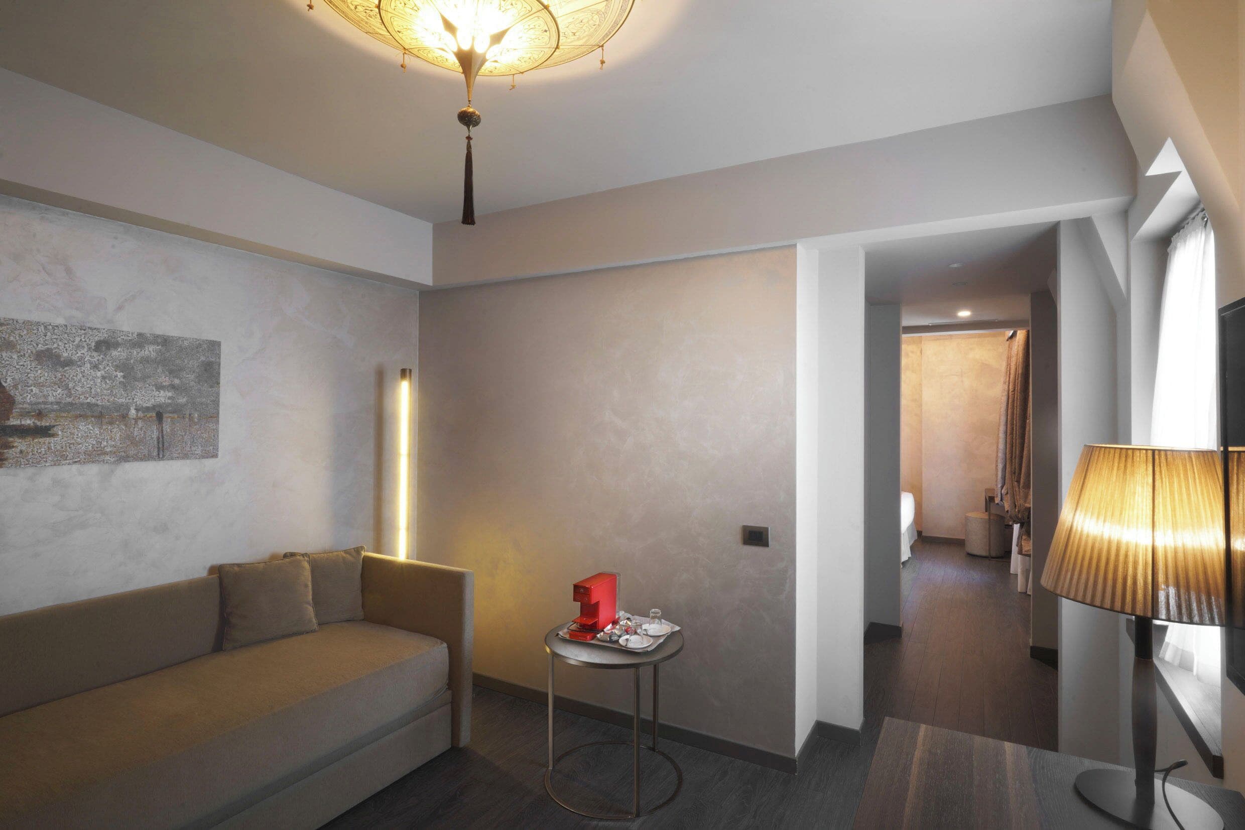 Moderni hotellihuone, jossa on sohva, pieni pöytä, jonka päällä on punainen esine, lamppu ja käytävä, joka johtaa kylpyhuoneeseen.