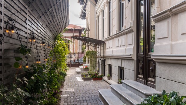 Une ruelle étroite pavée de pierres, avec une clôture en bois d’un côté et un bâtiment à la façade blanche et aux portes noires de l’autre.
