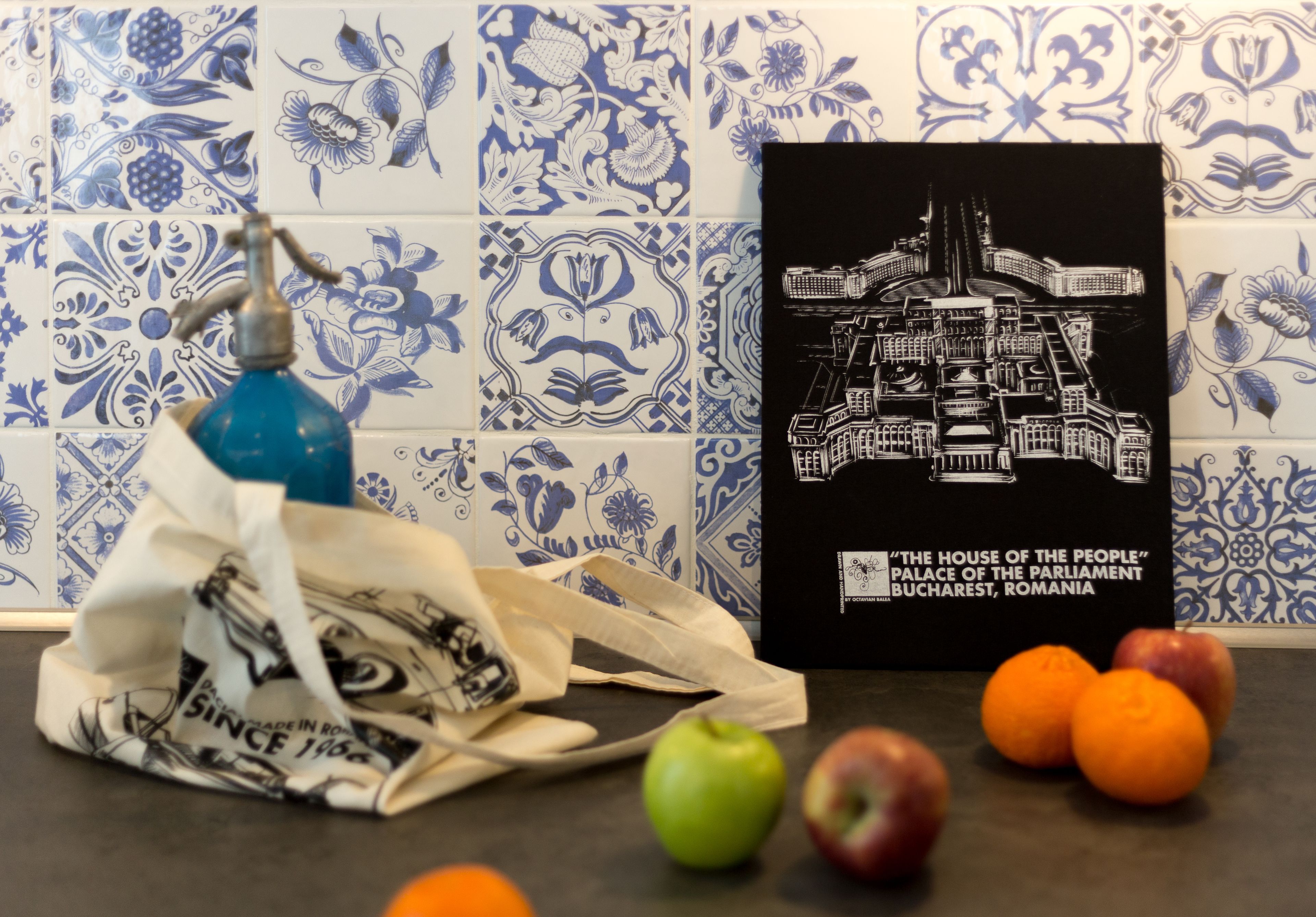 Una encimera de cocina con un salpicadero decorado con azulejos, un grabado blanco y negro del Parlamento, un grifo de agua azul, una bolsa de tela blanca y varias manzanas y naranjas.