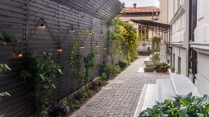 Un étroit sentier pavé de pierres, bordé d’une clôture en bois, de lampes suspendues et d’une végétation luxuriante.
