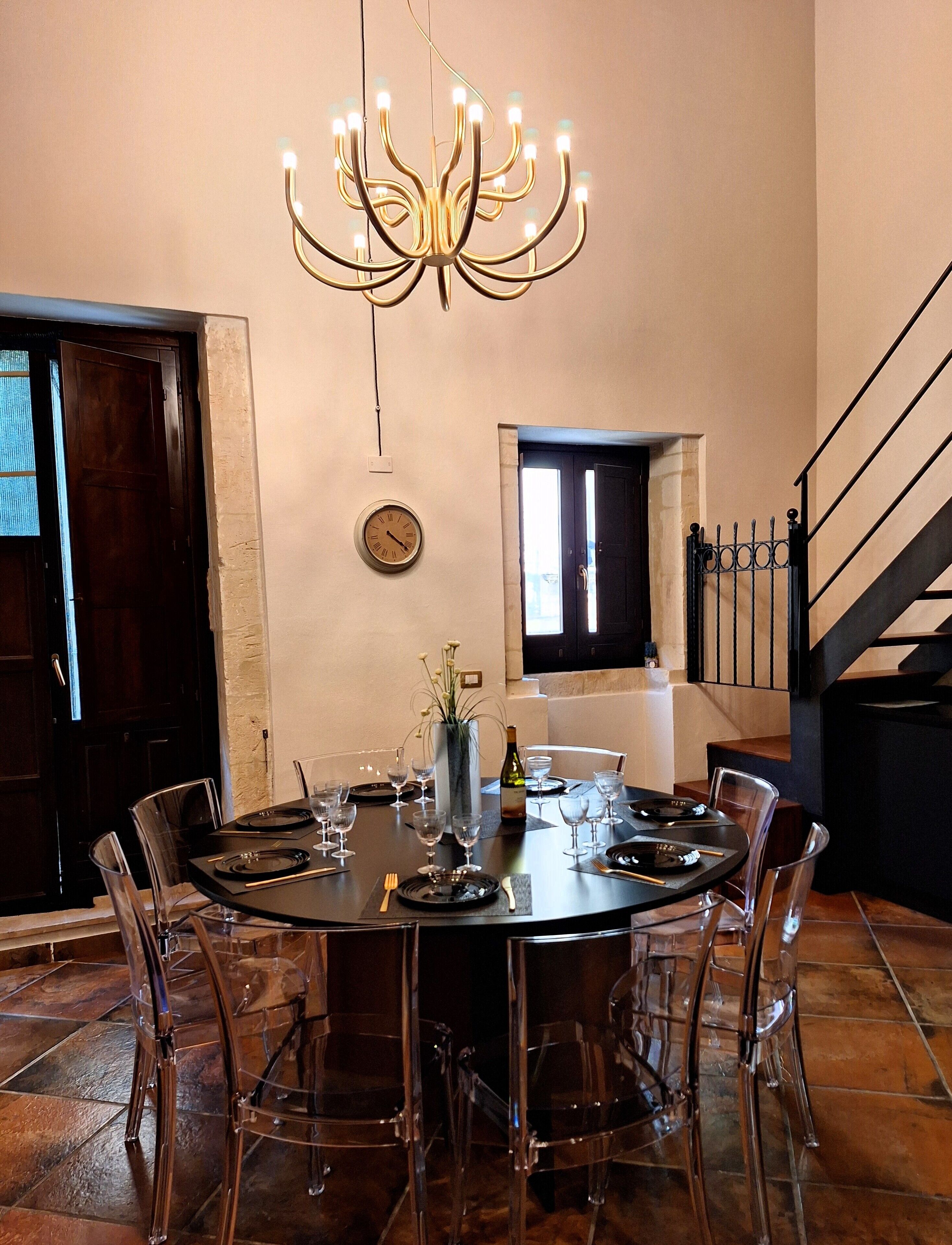 Un coin repas avec une table ronde dressée pour six personnes, entourée de chaises transparentes, d’un lustre, d’une horloge et d’une bouteille de vin.