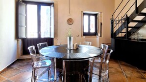 A dining area with a round table, clear chairs, a chandelier, and a staircase. - Casa Aretusa (Syracuse)