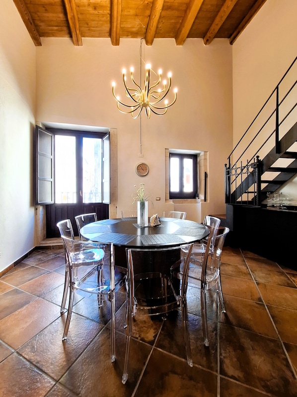 A dining area with a round table, clear chairs, a chandelier, and a staircase.