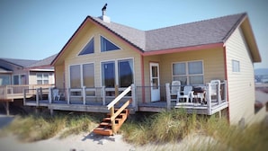 Een strandhuis met een houten terras, grote ramen en een trap naar de ingang.