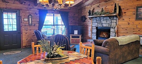 Acogedor interior de cabaña de troncos con chimenea de piedra, muebles de madera y un gran ventanal con cortinas.