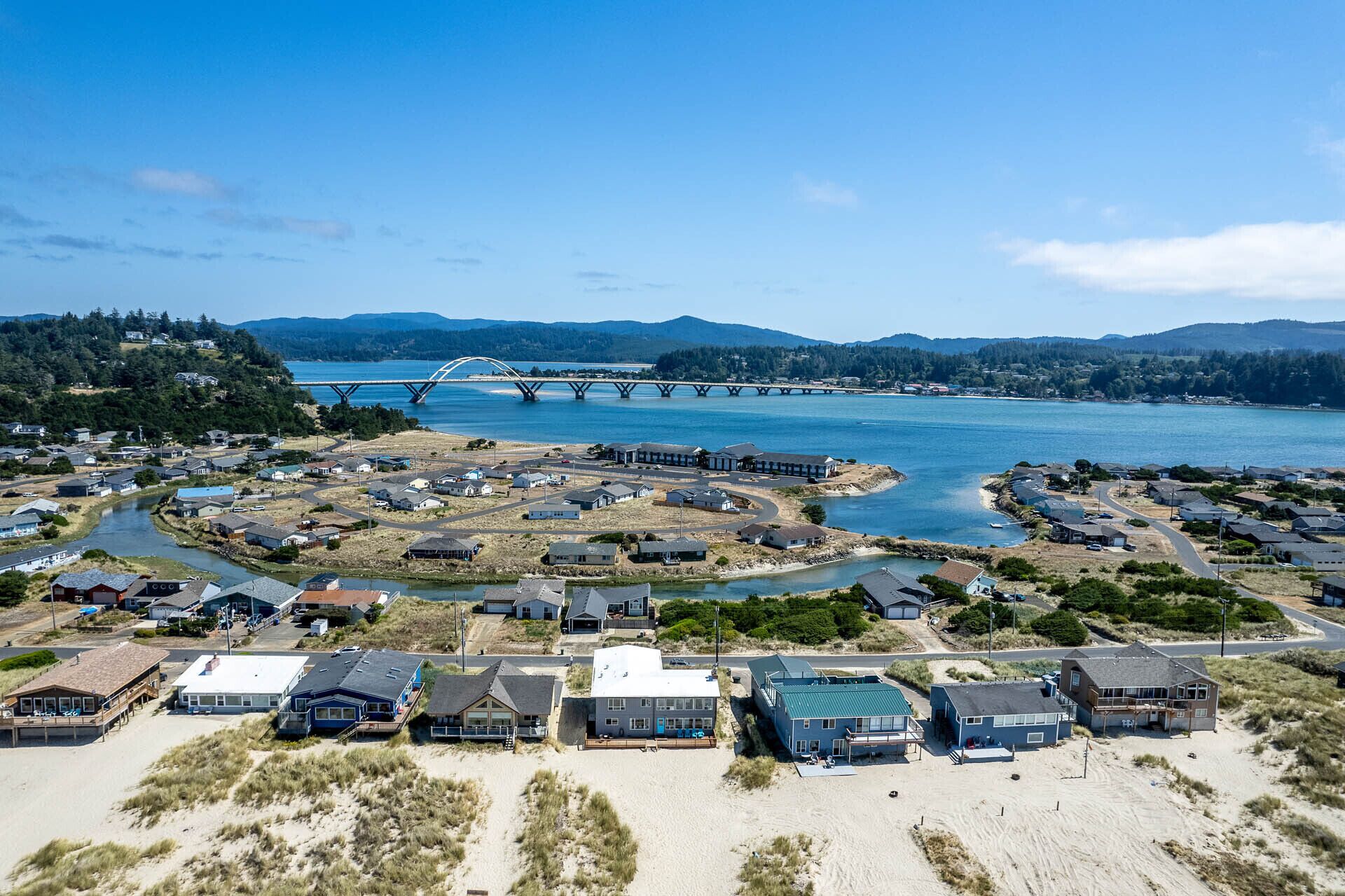 An aerial view of a coastal residential area with houses, a beach, and a bridge over a river.