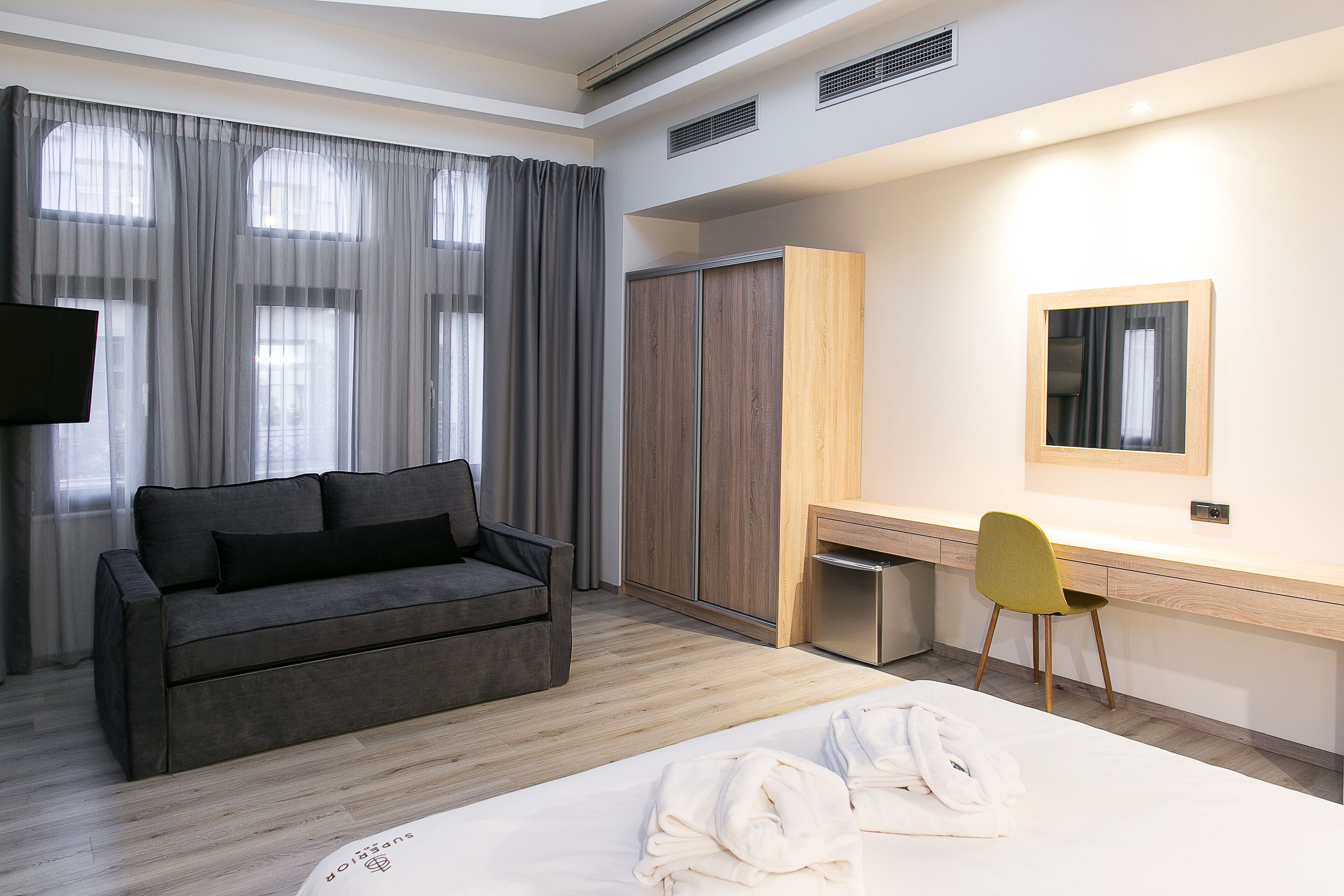 Una habitación de hotel moderna con un sofá gris, un escritorio de madera, una silla, una cama con ropa blanca y un armario.