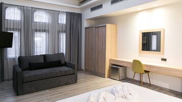 Una habitación de hotel moderna con un sofá gris, un escritorio de madera, una silla, una cama con ropa blanca y un armario.