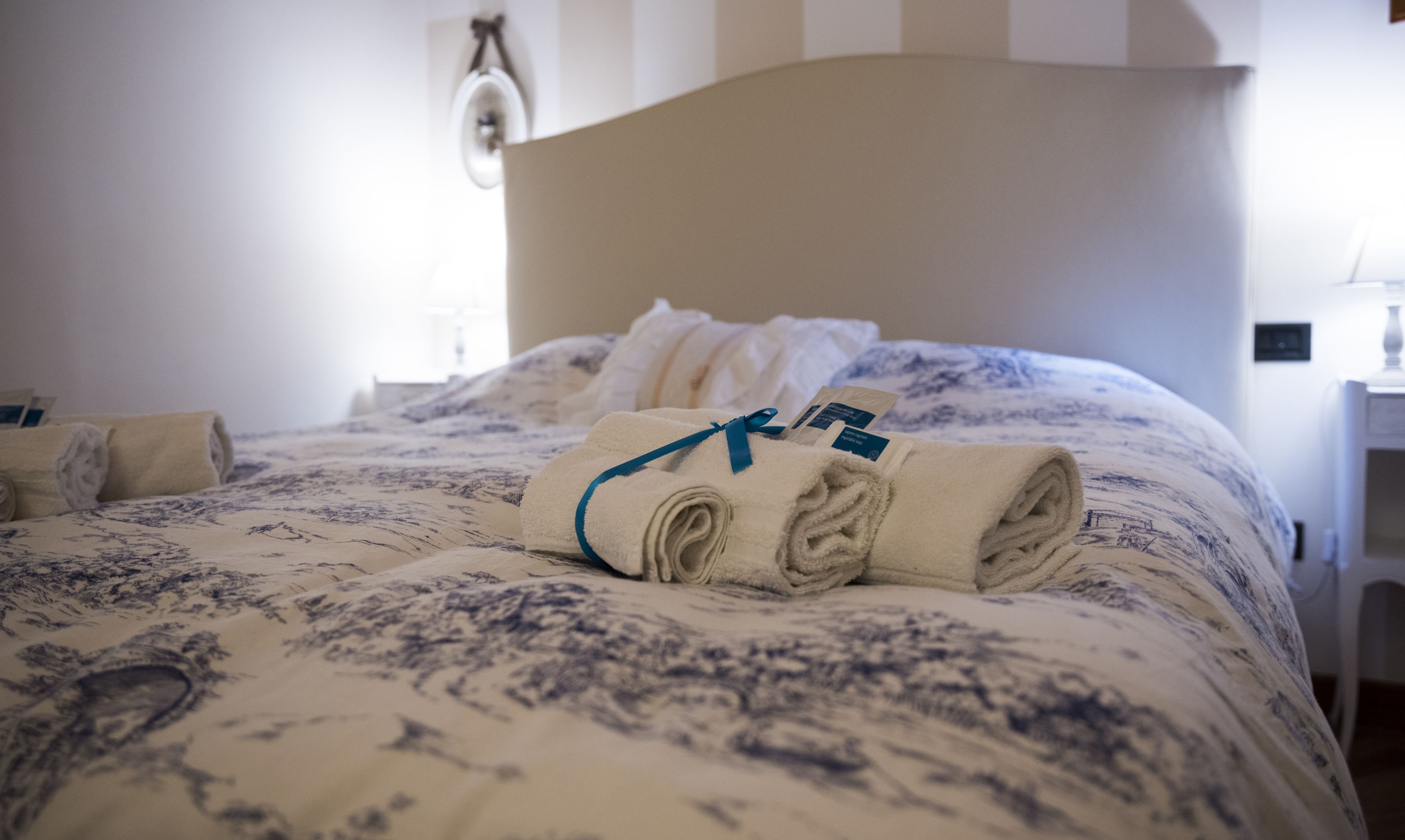Un lit bien fait, avec des serviettes pliées et un ruban bleu, une table de chevet avec une lampe et un luminaire fixé au mur.