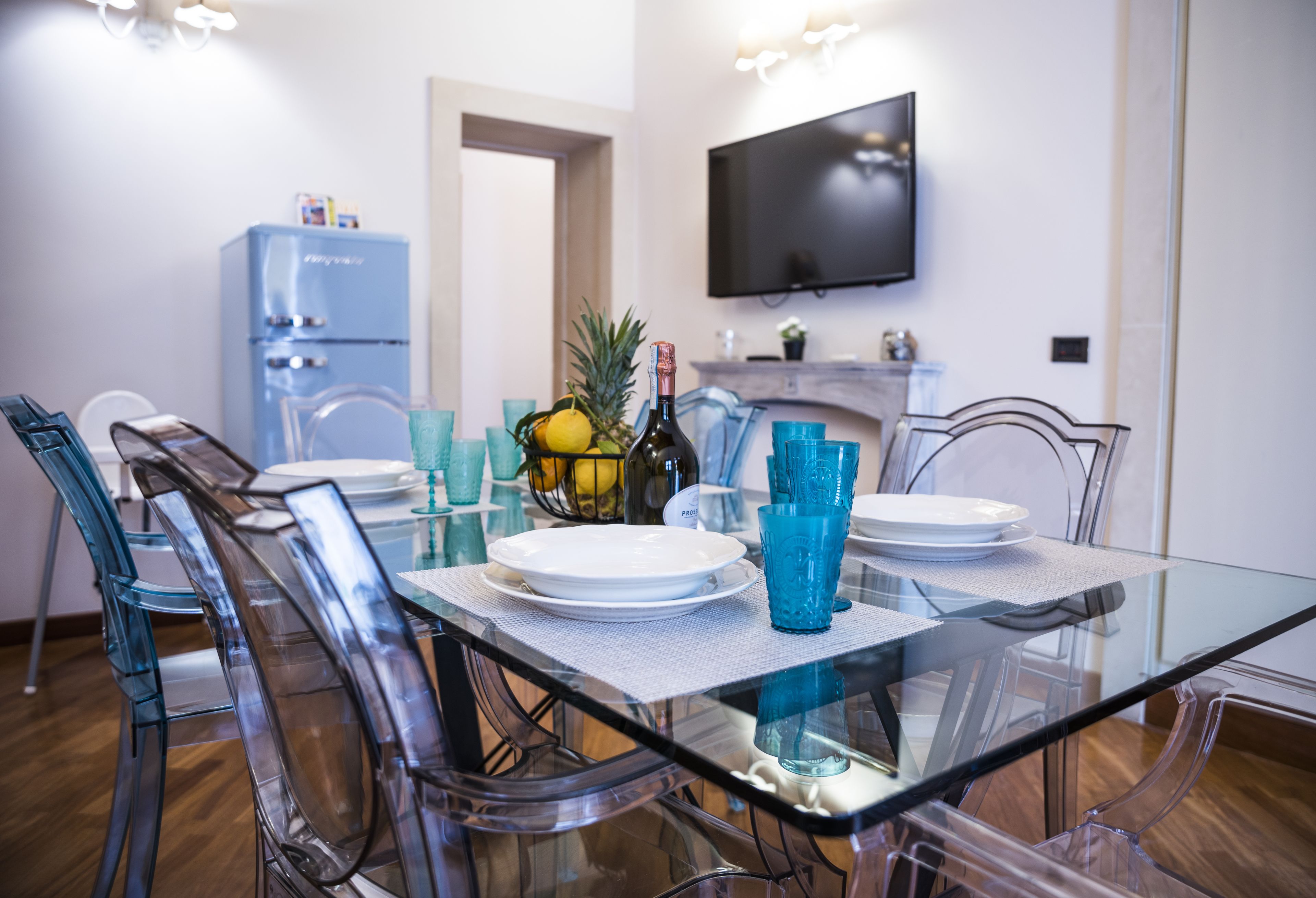 Un coin repas avec une table en verre, des chaises transparentes et un réfrigérateur bleu en arrière-plan.