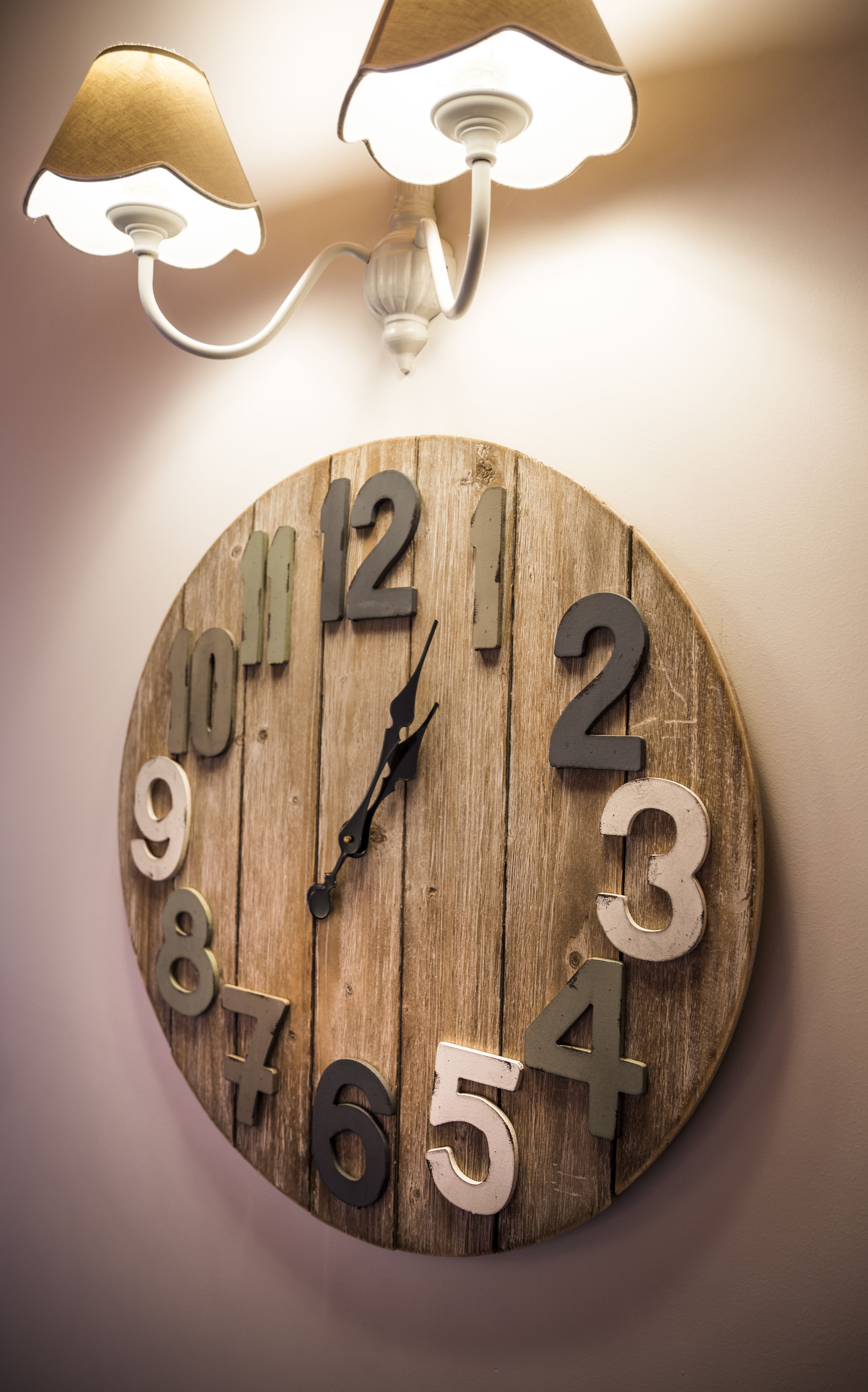 Un reloj de madera rústico con números y manecillas, colgado en una pared donde hay dos lámparas de pared arriba.