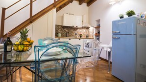 Une cuisine moderne avec des poutres en bois, un réfrigérateur bleu et un coin repas avec des chaises et une table en verre transparent.