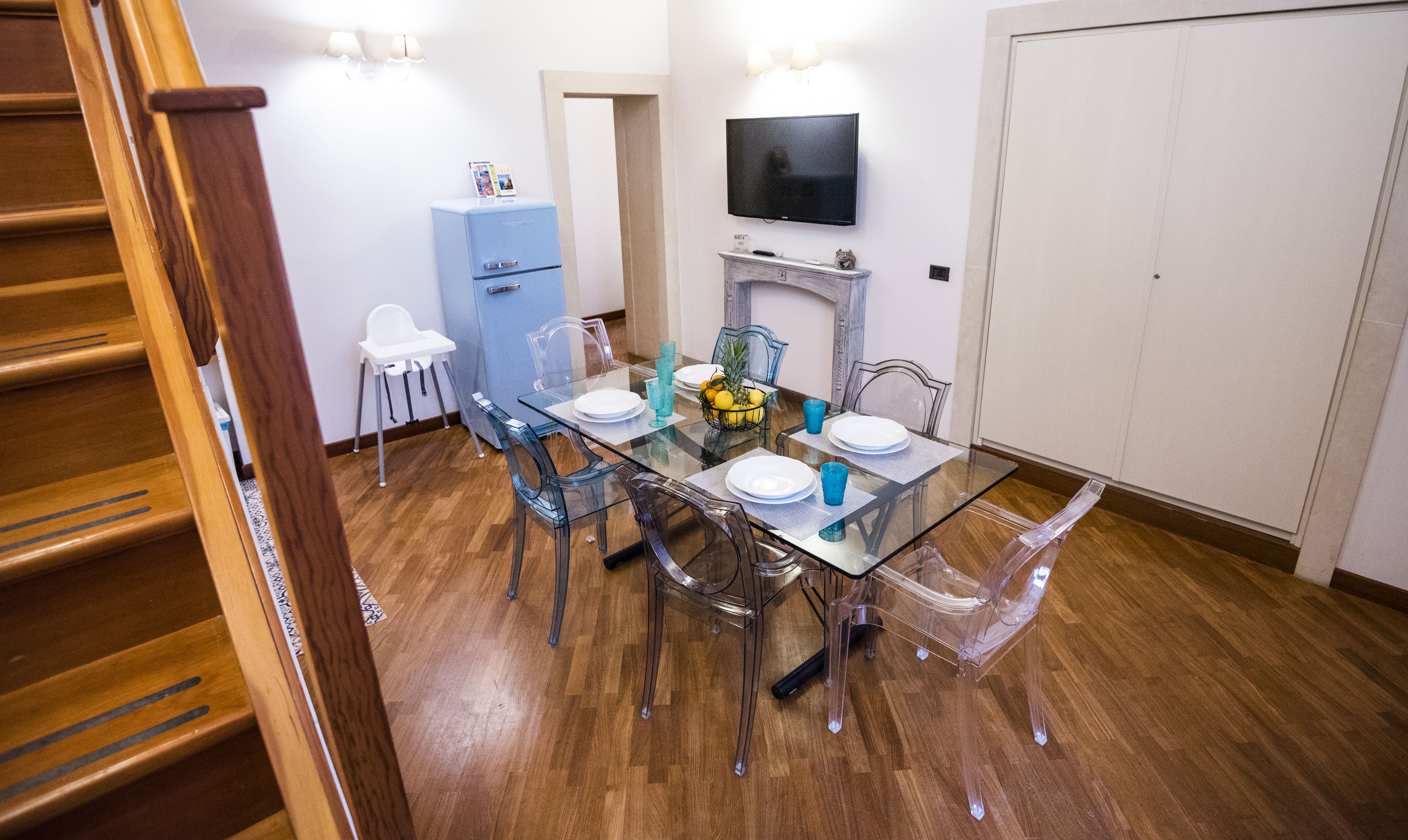 A dining area with a glass table, clear chairs, a blue refrigerator, and a TV mounted on the wall.
