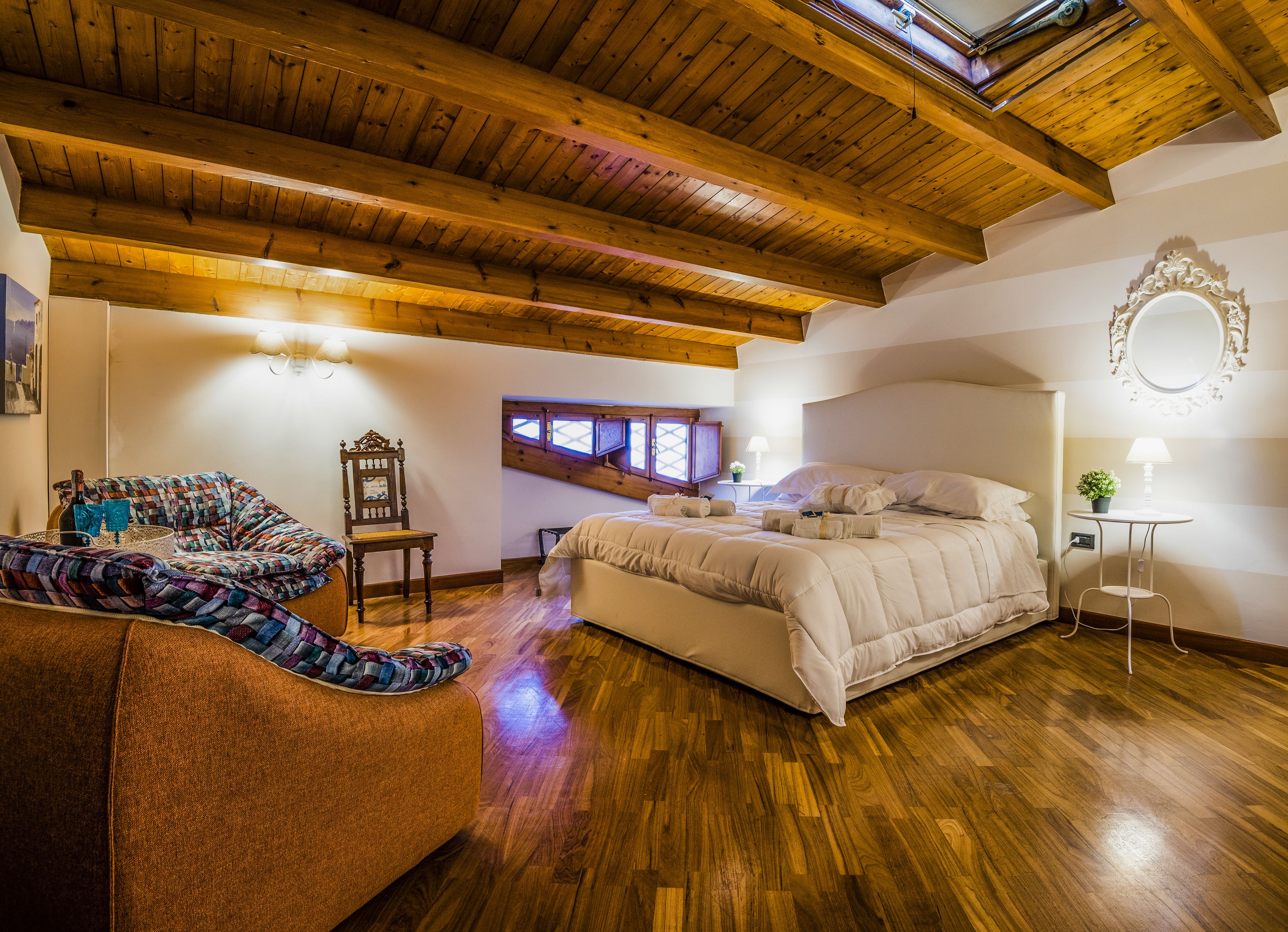 Camera da letto con soffitto in legno, un letto, una sedia, un comodino con una lampada e una finestra vista.