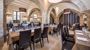 Un restaurante con arcos de piedra, varias mesas dispuestas con copas de vino y cubiertos, y una estantería con botellas de vino al fondo.