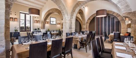 A restaurant with stone arches, multiple dining tables set with wine glasses and cutlery, and a wine rack in the background.