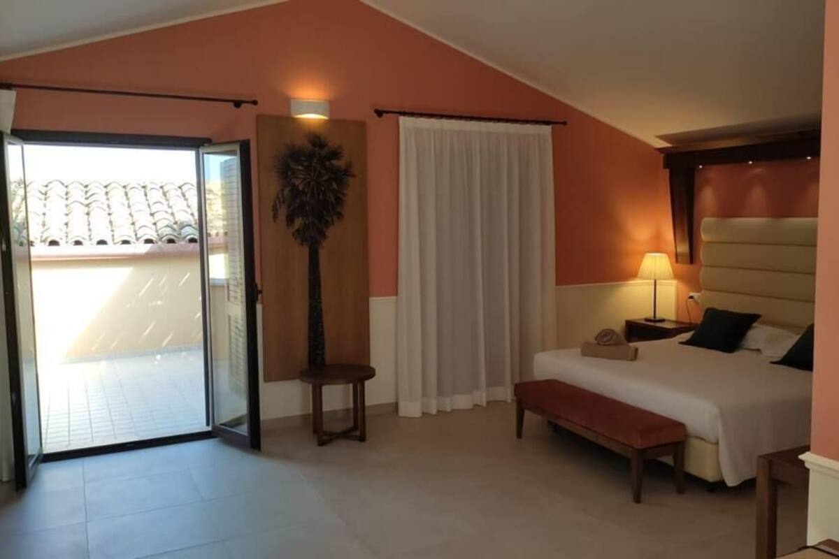 Una camera d'albergo con un letto, una panca, un comodino, una lampada, una pianta e una porta scorrevole in vetro che dà su un balcone.
