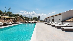 Una piscina rectangular con sillas de descanso y sombrillas a ambos lados, rodeada por un área ajardinada con árboles y un edificio de techo de tejas.