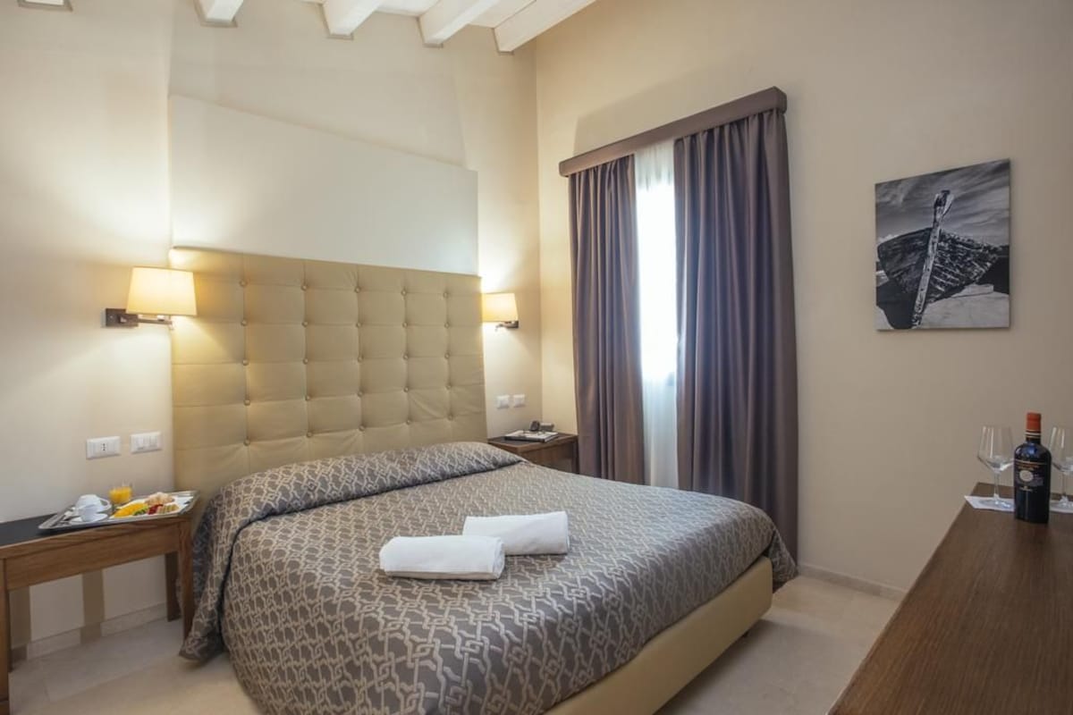 A hotel room with a large bed, a bedside table with a lamp, a nightstand, a window with curtains, and a painting on the wall.