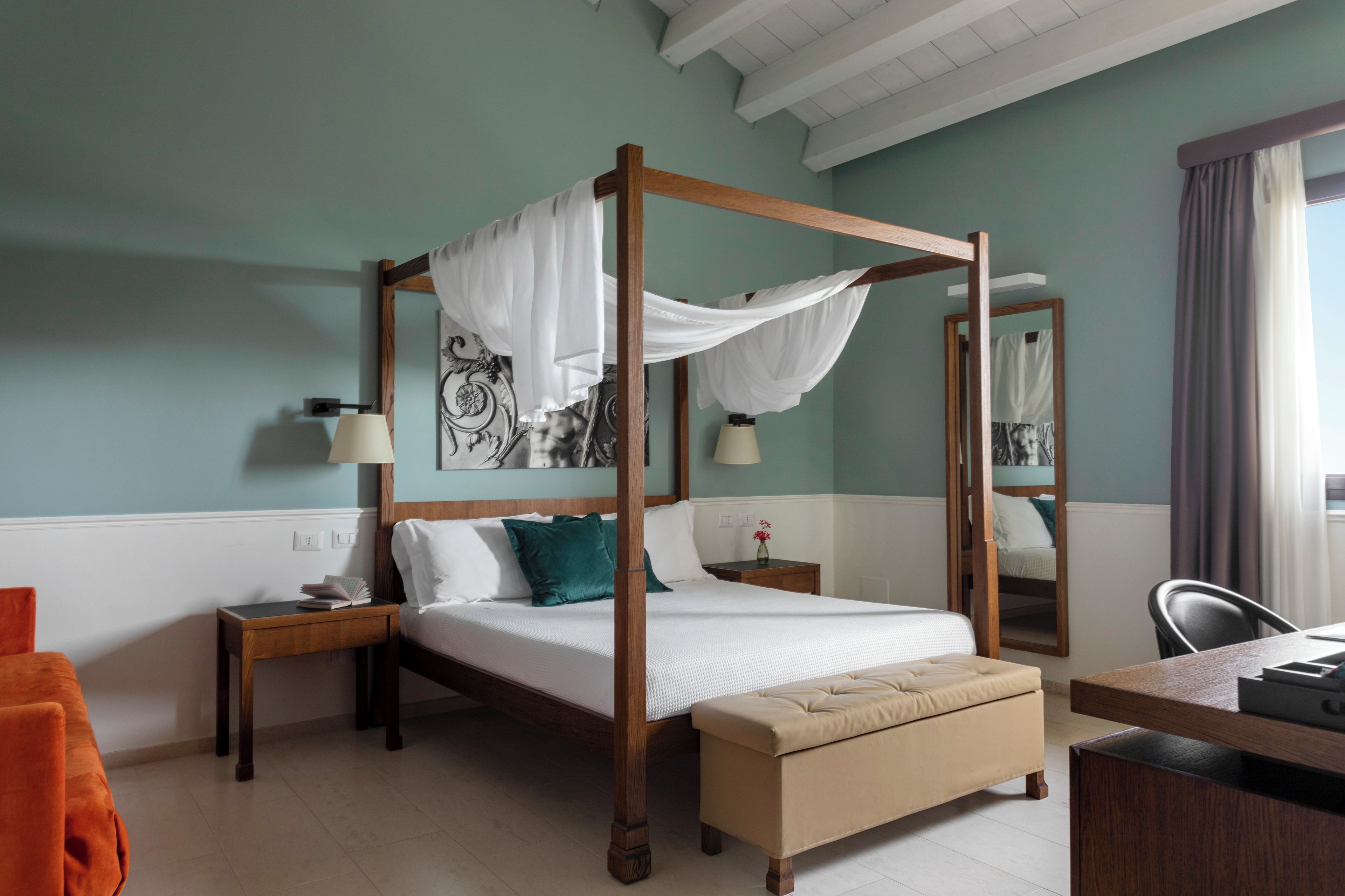 A four-poster bed with a canopy, a bedside table with a lamp, a small bedside table with a vase, a bench at the foot of the bed, and a chair.