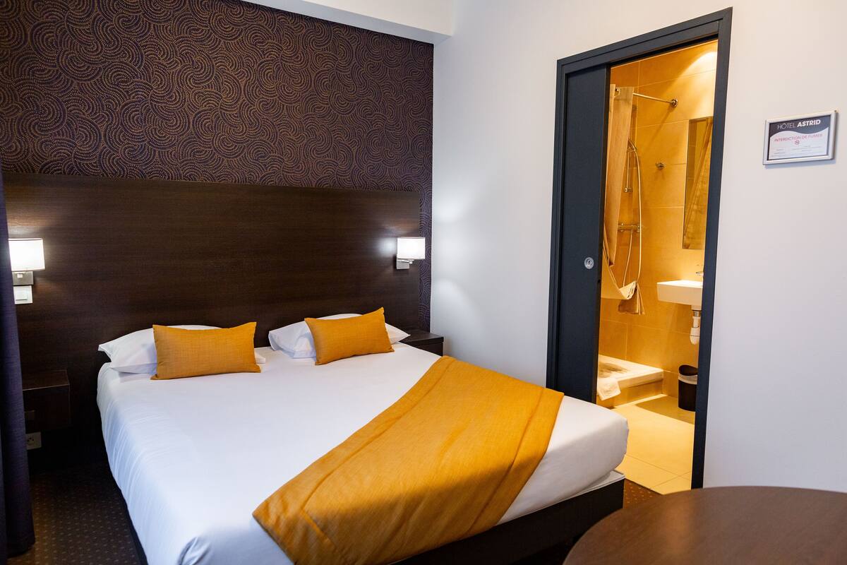A hotel room with a large bed, two bedside lamps, a wooden headboard, and a bathroom visible through an open door.