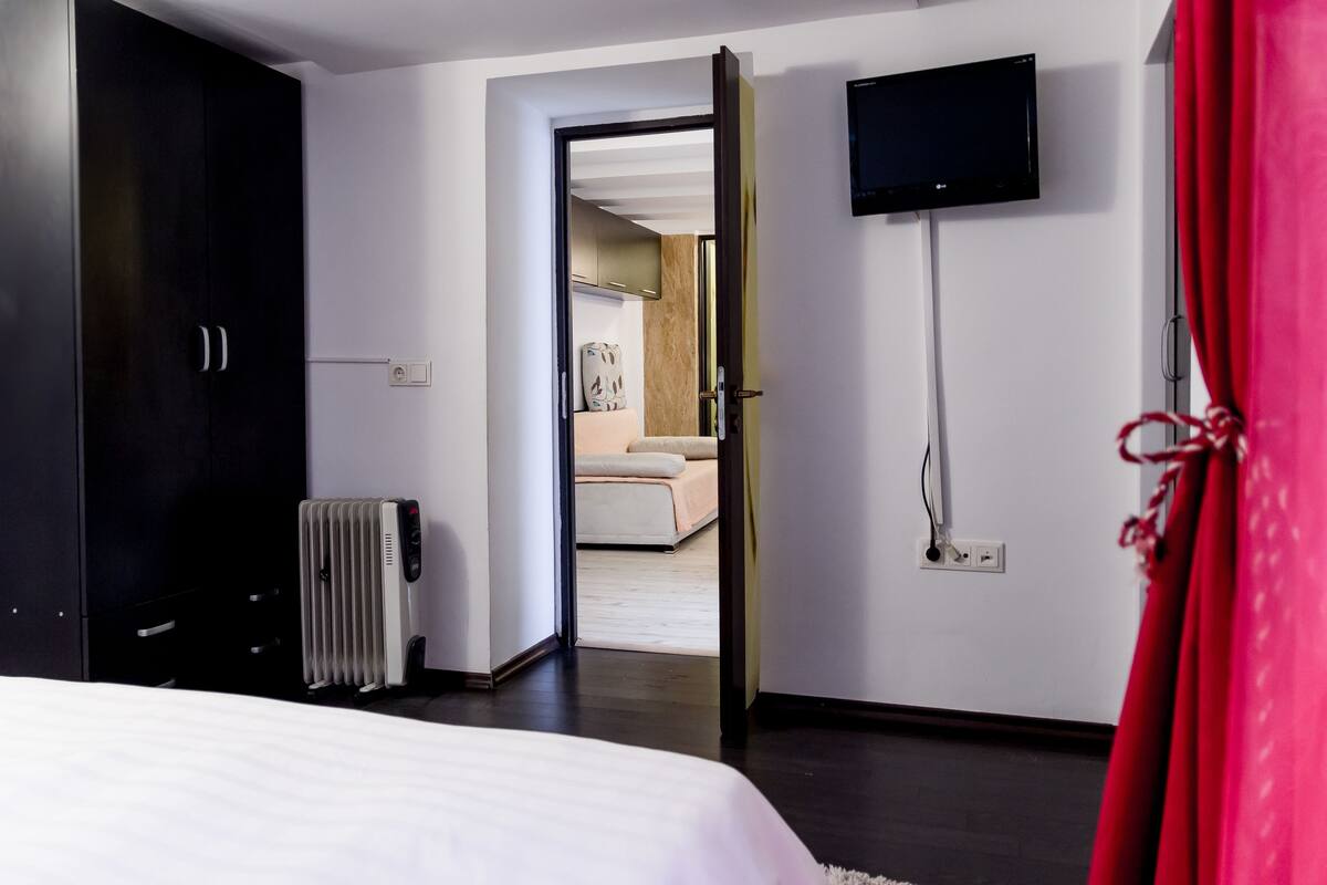 A modern hotel room with a bed, a radiator, a TV mounted on the wall, and a view into the adjacent room.