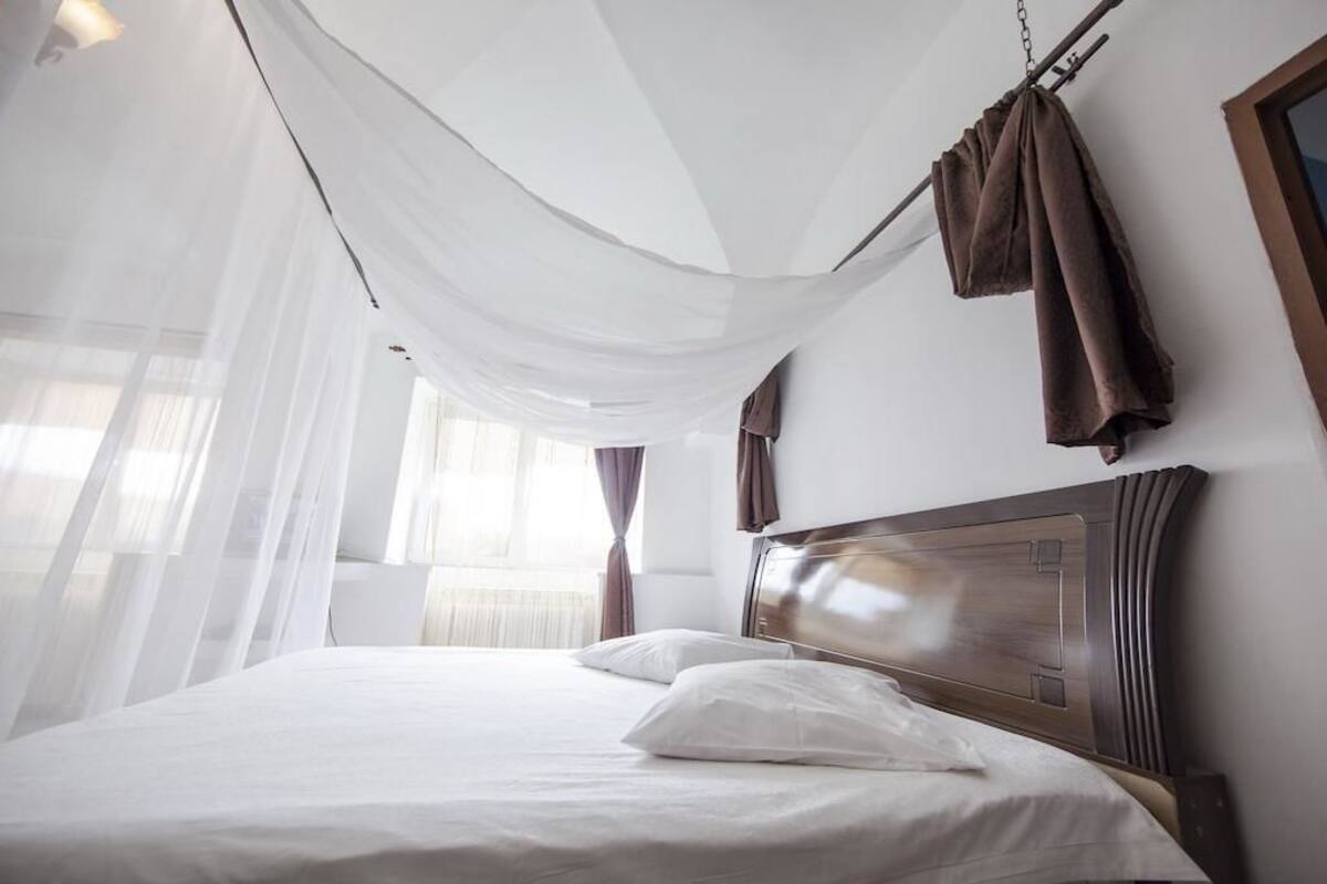 A hotel room with a bed, white curtains, and a wooden headboard.