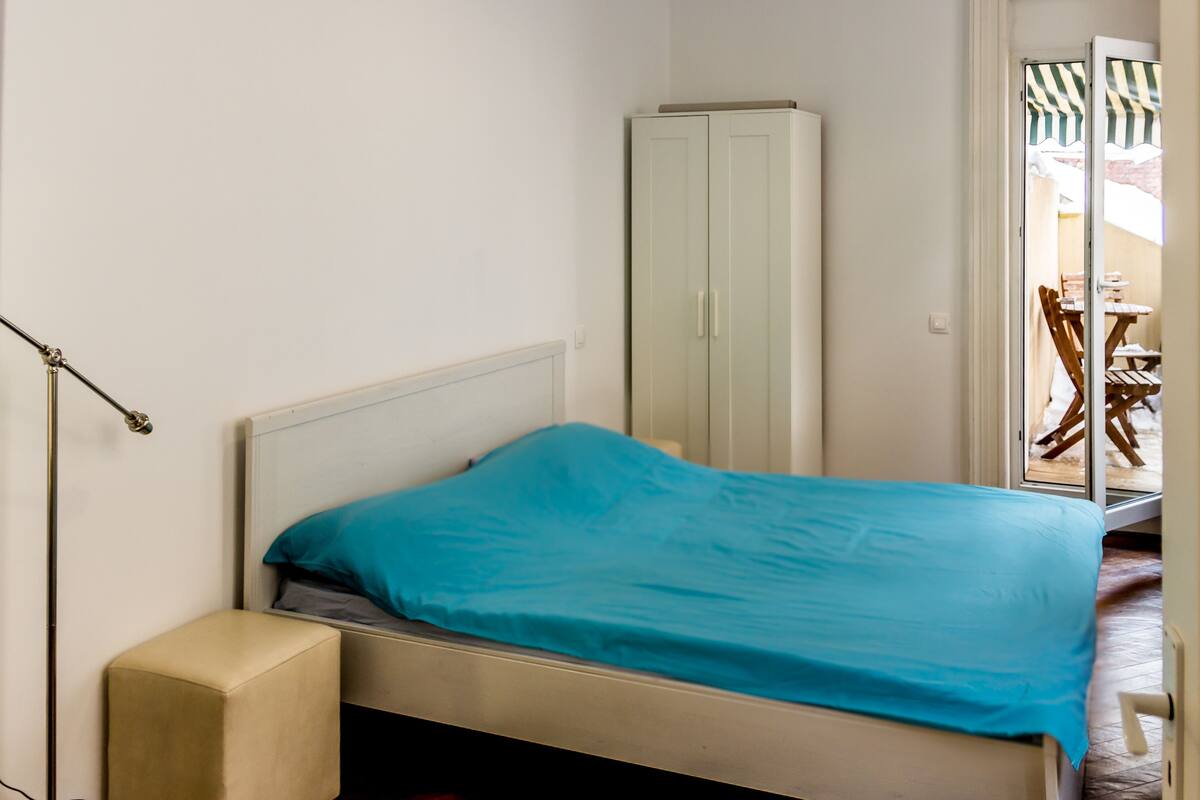A small bedroom with a single bed, a wardrobe, and a view of an outdoor seating area.