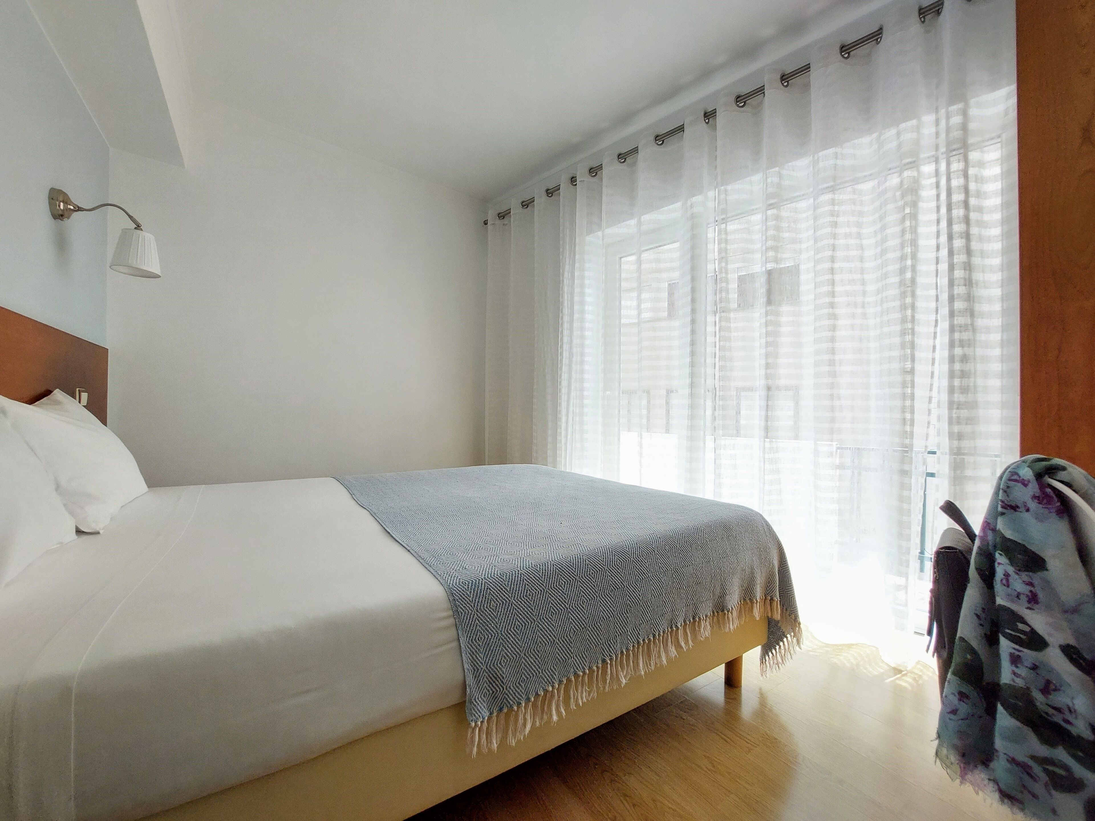 Habitación de hotel con cama, cabecera, lámpara de noche, ventana con cortinas translúcidas y suelo de madera.