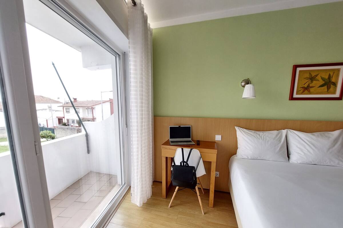 A hotel room with a bed, a desk with a laptop, a chair, and a window with a view of buildings.
