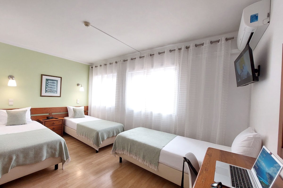 A hotel room with two single beds, a desk with a laptop, a TV, and a window with sheer curtains.