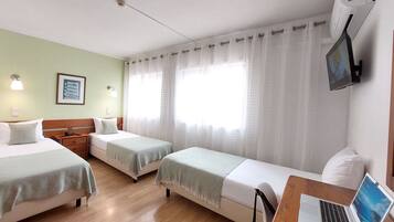 A hotel room with two single beds, a desk with a laptop, a TV, and a window with sheer curtains.