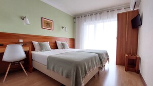 Habitación de hotel con cabecera de madera, dos camas con mantas de estampado verde, una silla blanca, un taburete de madera pequeño, un televisor montado en la pared y una ventana con cortinas traslúcidas.