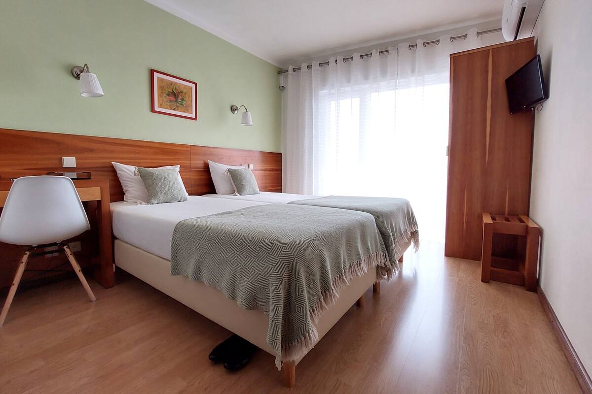A hotel room with a wooden headboard, two beds with green patterned blankets, a white chair, a small wooden stool, a TV mounted on the wall, and a window with sheer curtains.