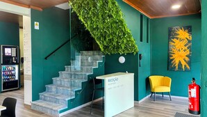 Un mostrador de recepción con un cartel que indica el horario, una escalera con pared verde y una planta, una silla amarilla y un extintor.