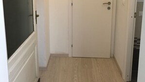 Un couloir étroit, avec une porte blanche au fond, une porte avec un panneau de verre noir sur le côté gauche, et une pièce partiellement visible dont le sol est carrelé.